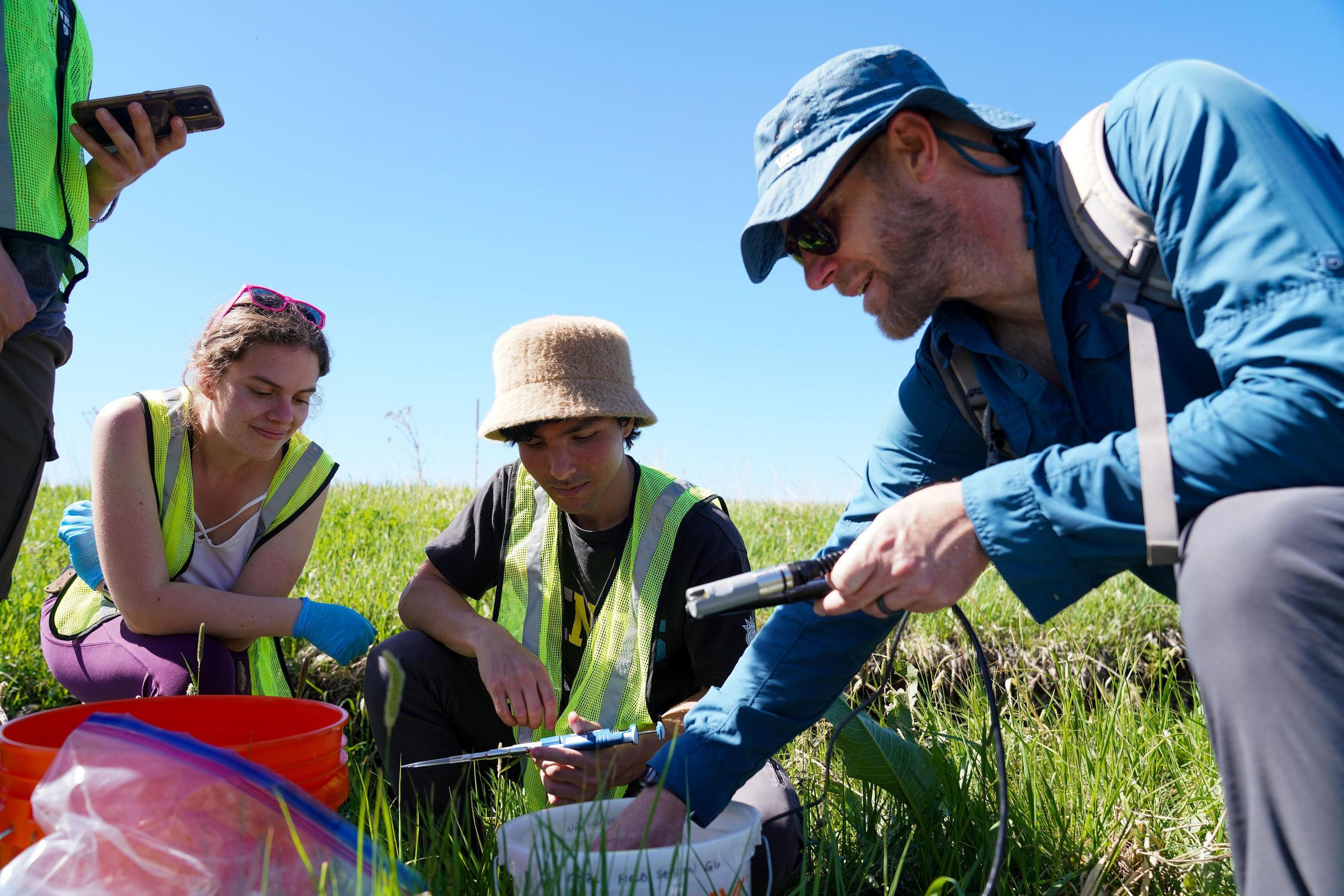 Field researchers in safety vests examine samples and equipment in a grassy field under a clear blue sky.